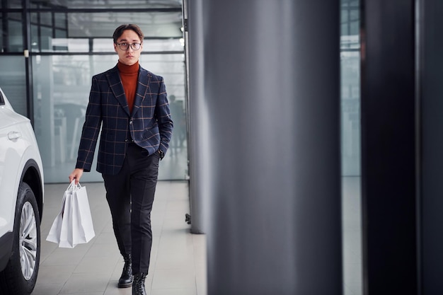 Tenant des sacs à provisions. Jeune homme d'affaires en costume de luxe et vêtements formels est à l'intérieur près de la voiture.