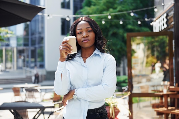 Tenant une tasse de boisson dans les mains Jeune femme afro-américaine en chemise blanche à l'extérieur de la ville près des arbres verts et contre le bâtiment de l'entreprise