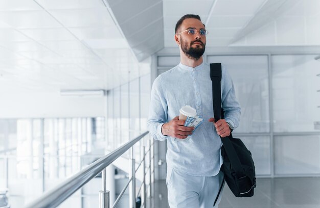 Tenant une tasse avec boisson et sac noir Jeune bel homme en vêtements formels à l'intérieur dans le bureau pendant la journée