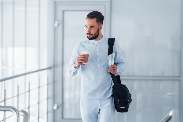 Tenant une tasse avec boisson et sac noir Jeune bel homme en vêtements formels à l'intérieur dans le bureau pendant la journée