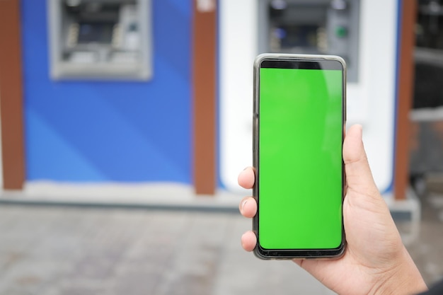 Tenant un téléphone intelligent avec écran vert sur le fond de l'ATM