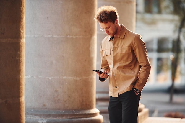 Tenant le téléphone à la main Jeune homme élégant dans des vêtements chics formels à l'extérieur de la ville