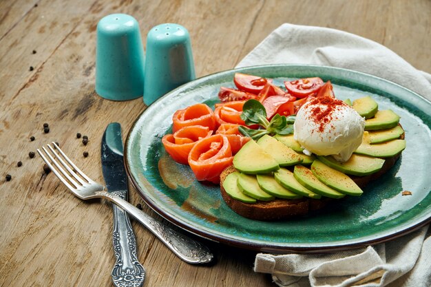 Tendance alimentaire - toast à l'avocat avec œuf poché sur pain de seigle avec saumon sidedish, tomate dans une assiette en céramique sur une surface en bois. Petit déjeuner sain