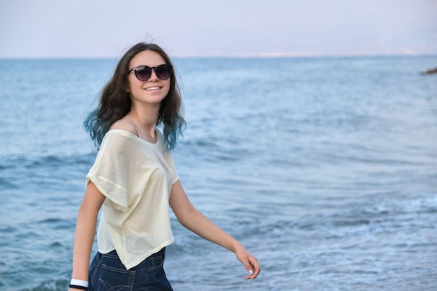 Tendance belle fille adolescente avec de longs cheveux bleus colorés en lunettes de soleil et shorts marchant le long de la plage