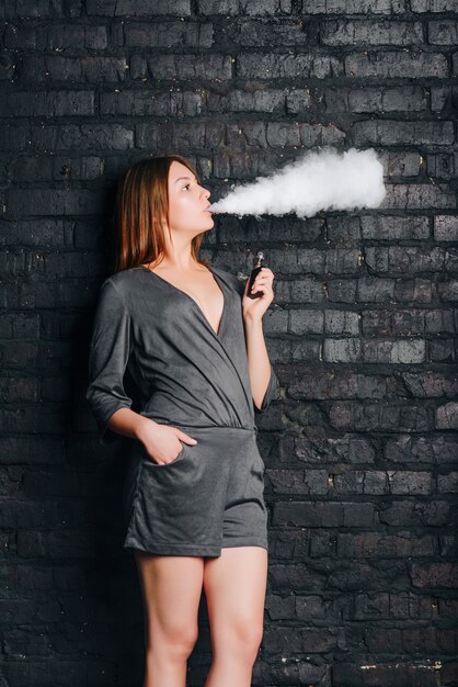 Tendance belle fille fumant une cigarette sans tabac, exhalant de gros nuages de fumée. Vêtu de vêtements à la mode.