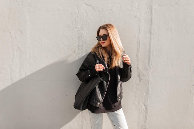 Tendance jeune belle femme avec des lunettes de soleil en veste à capuche en cuir cool et un jean avec un sac en cuir noir se tient près d'un mur gris dans la rue