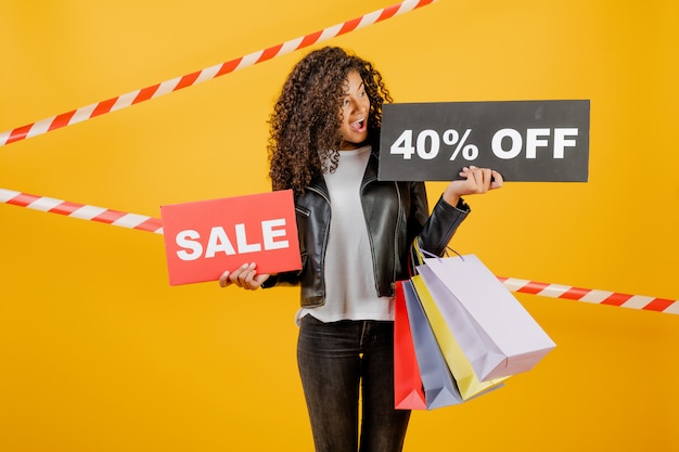 Photo tendance jeune fille noire avec signe de vente 40% et sacs colorés isolés sur jaune avec du ruban de signalisation