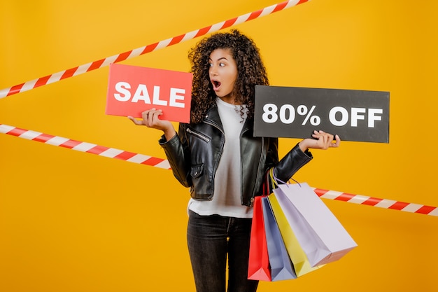 Photo tendance jeune fille noire avec signe de vente 80% et sacs colorés isolés sur jaune avec du ruban de signalisation