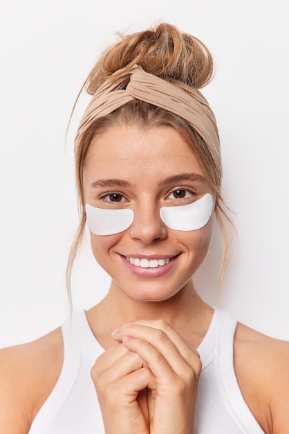 Tendre charmante jeune femme sourit doucement garde les mains ensemble vêtue d'un t-shirt décontracté applique des patchs de beauté sous les yeux pour les poses de traitement de la peau sur fond blanc. Masque de rajeunissement