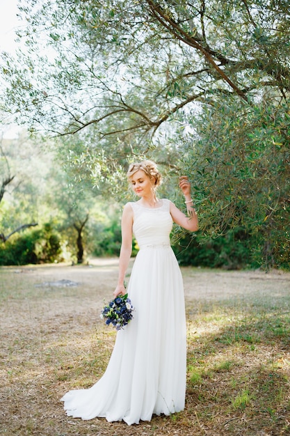 Une tendre mariée avec un bouquet de fleurs bleues se tient près d'un olivier et touche les branches avec