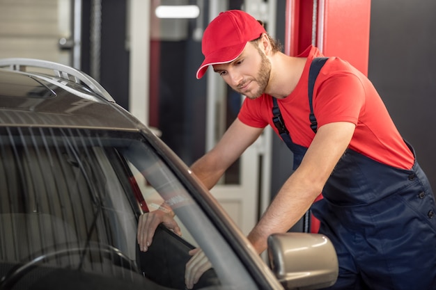 Étendue des travaux. Intéressé jeune homme barbu en vêtements de travail furtivement dans la fenêtre de la voiture ouverte en atelier