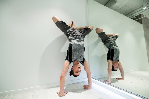 Tenez-vous près du mur. Homme aux cheveux noirs portant un short gris et un t-shirt faisant un bras près du mur et du miroir