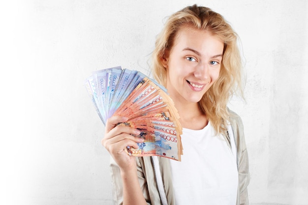 Tenge. Une belle fille blonde dans un t-shirt blanc sourit et tient dans sa main beaucoup de factures kazakhes sur un gros plan de fond blanc.