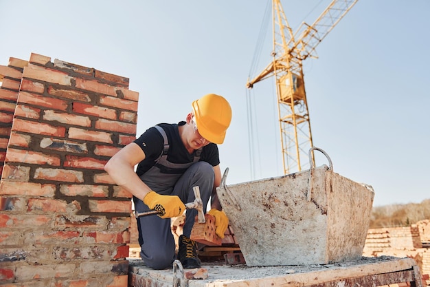 Tenir la brique et utiliser un marteau Un ouvrier du bâtiment en uniforme et équipement de sécurité a un travail sur la construction