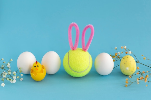 Tennis de Pâques avec balle de tennis smiley avec oreilles de lapin, œufs de poule blancs et gypsophile sur fond bleu.