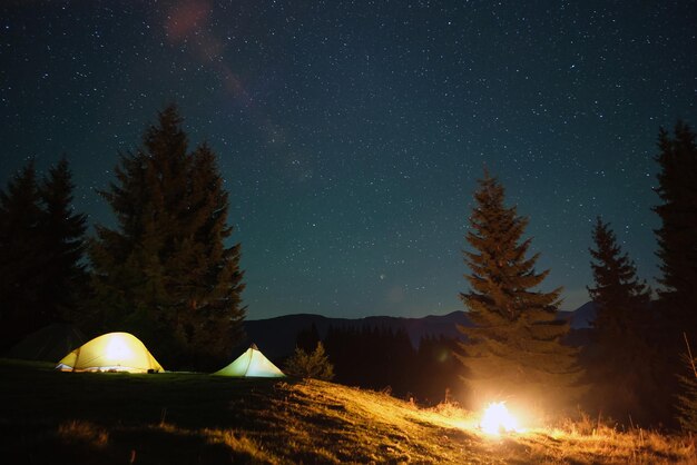 Tentes touristiques lumineuses illuminées près d'un feu de joie rougeoyant sur un site de camping dans des bois de montagne sombres sous un ciel nocturne avec des étoiles scintillantes Mode de vie actif et concept de vie en plein air