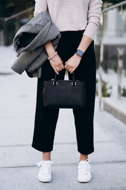 Tenue élégante. Gros plan du grand sac noir texturé. Modèle posant dans la rue, portant un pantalon court, un pull crémeux, un manteau ou une veste gris et des baskets blanches. Concept de mode féminine.