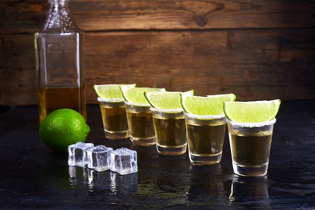 Tequila mexicaine en or dans des verres courts avec du sel, des tranches de citron vert et de la glace sur une table en bois. Fumée.
