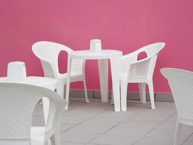 Terrasse de café en plein air avec mur rose, chaises et tables en plastique blanc.