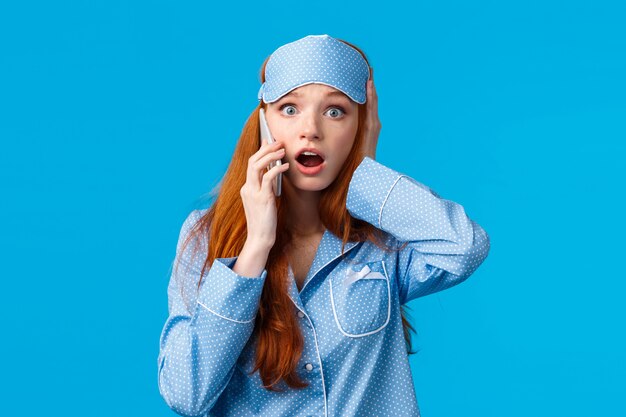 Terribles nouvelles. Choquée et inquiète jeune fille troublée entendant une mauvaise chose arriver, parler au téléphone, toucher la tête frustrée, haletant la mâchoire et regarder stupéfait, mur bleu debout