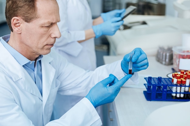 Test sanguin. Photo de profil d'un praticien compétent tout en tenant un verre de test avec du sang et en allant le tester assis dans le laboratoire