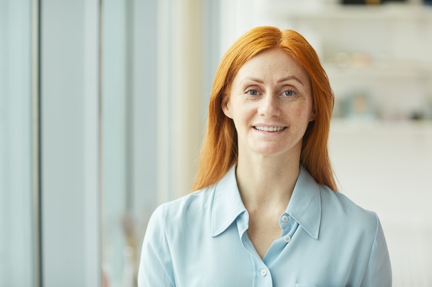 Tête et épaules portrait de femme d'affaires aux cheveux rouges souriant en se tenant debout par fenêtre dans un bureau blanc moderne, espace copie