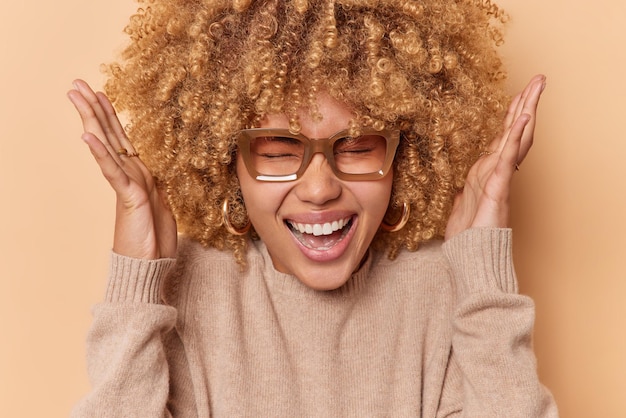 La tête d'une femme bouclée heureuse et ravie lève les mains sur la tête et s'exclame joyeusement porte des lunettes et des poses de pull en cachemire sur fond marron réagit à des nouvelles incroyables. Notion d'émotions