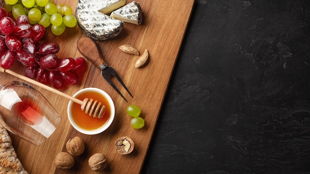 Tête de fromage, grappe de raisin, miel, noix et verre à vin sur planche de bois et fond noir avec fond