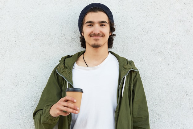 Tête d'homme hipster heureux avec du café à emporter, vêtu d'un t-shirt blanc et d'une veste verte, aime le temps libre, porte un chapeau noir, des mannequins sur fond blanc, a les cheveux bouclés. Les gens et boire