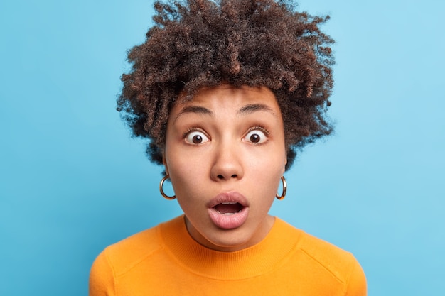 Photo la tête d'une jeune femme afro-américaine choquée regarde avec les yeux largement ouverts et la bouche réagit sur des stands de nouvelles incroyables dans la stupeur porte un pull orange décontracté isolé sur un mur bleu