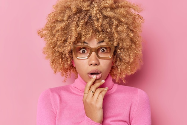 Tête d'une jeune femme aux cheveux bouclés impressionnée qui a l'air effrayée par la caméra garde la bouche ouverte a retenu son souffle ne peut pas croire que ses yeux portent des lunettes et un col roulé isolé sur fond rose