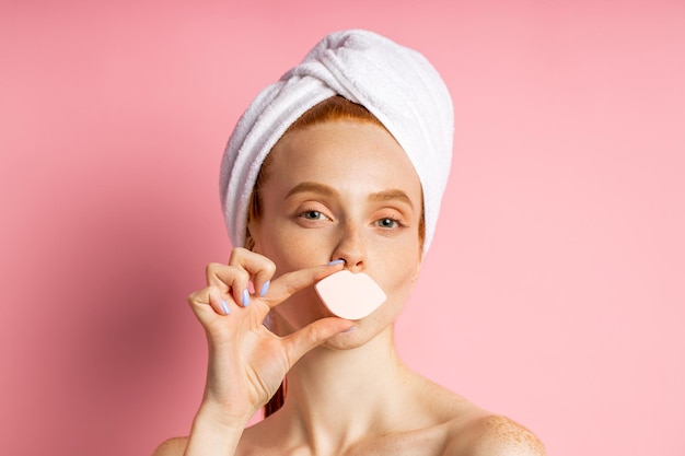 Tête d'une jeune femme gaie au gingembre avec une peau fraîche et propre, ayant des procédures d'hygiène, portant une serviette sur la tête, couvre la bouche d'une éponge cosmétique en forme de lèvres, posant sur fond rose.