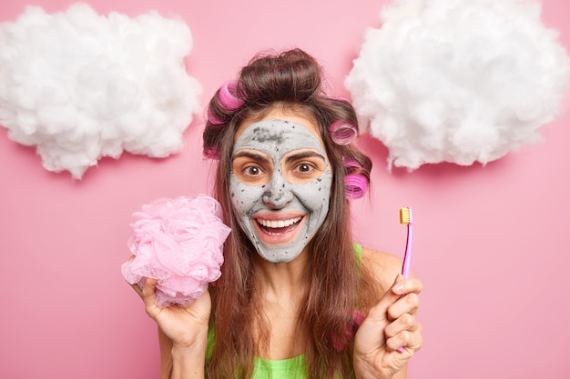 Tête de jolie femme européenne positive applique un masque d'argile sur les rouleaux de cheveux du visage tient une brosse à dents et une éponge de bain a des routines matinales isolées sur un mur rose