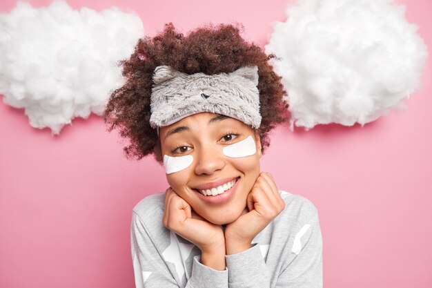 Tête de jolie femme souriante garde les mains sous le menton regarde joyeusement la caméra porte un masque de sommeil et un pyjama bénéficie d'un bonjour applique des patchs de beauté sous les yeux isolés sur un mur de studio rose