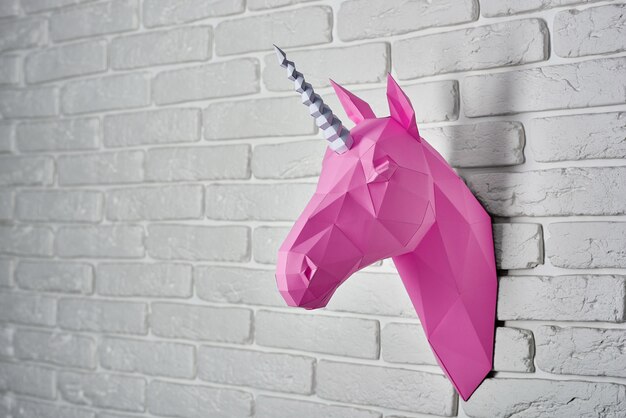 Tête de licorne rose saturée, faite de papier accroché sur un mur de briques blanches.