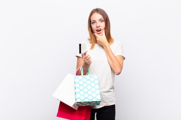 Tête rouge jolie femme avec la bouche et les yeux grands ouverts et la main sur le menton, se sentant désagréablement choqué, disant quoi ou wow avec des sacs à provisions