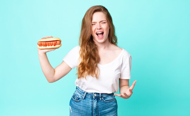 Tête rouge jolie femme à la colère, agacée et frustrée et tenant un hot-dog