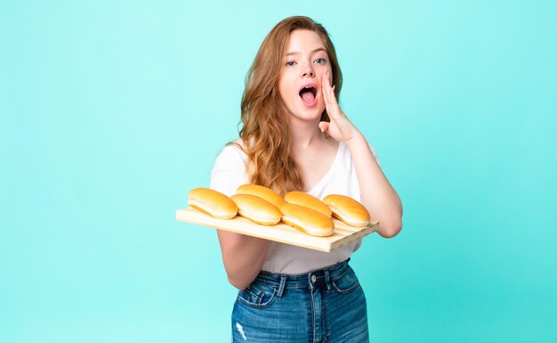 Tête rouge jolie femme se sentant heureuse en donnant un grand cri avec les mains à côté de la bouche et tenant un pain troy