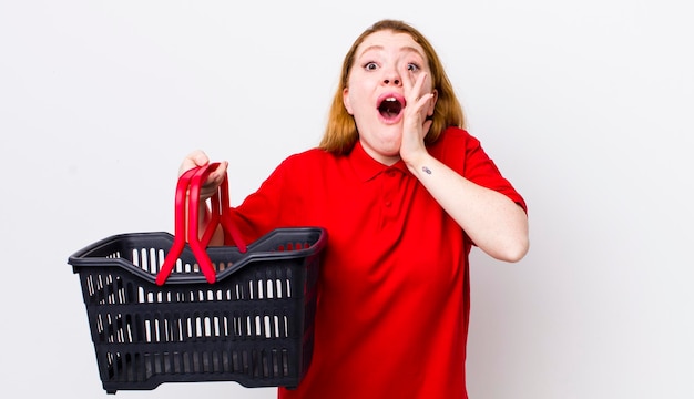 Tête rouge jolie femme se sentant heureuse de donner un grand cri avec les mains à côté de la bouche concept de panier vide