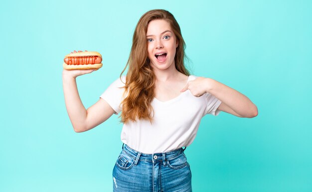 Tête rouge jolie femme se sentant heureuse et pointant vers soi avec un excité et tenant un hot-dog