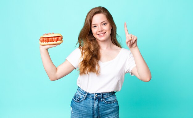 Tête rouge jolie femme souriante et semblant amicale, montrant le numéro un et tenant un hot-dog