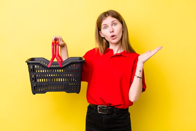 Tête rouge jolie femme à la surprise et choquée avec la mâchoire tombée tenant un concept de panier d'achat d'objet