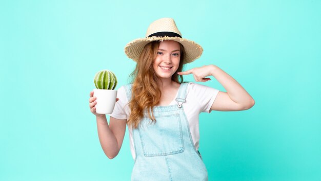 Tête rouge jolie fermière souriante pointant avec confiance vers son propre large sourire et tenant un cactus