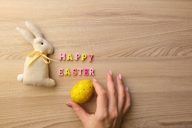 Texte en bois Joyeuses Pâques sur une table en bois, une main de femme avec un oeuf peint et un lapin fait à la main en tissu