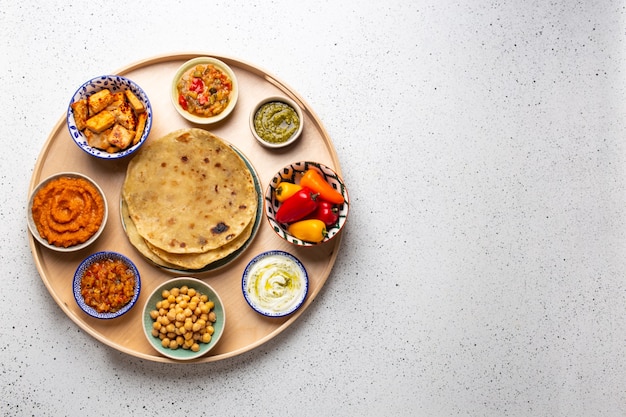 Thali indien - sélection de divers plats servis sur un plateau en bois rond, table blanche. Assortiment de mezes végétariens indiens avec pain chapati, paneer, dal, chutney dans des bols. Vue de dessus, espace pour le texte