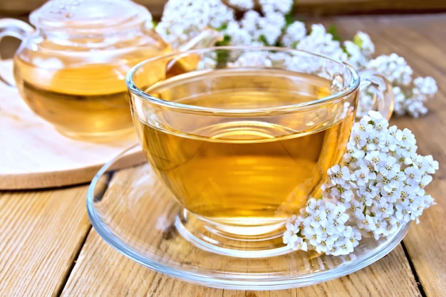 Thé d'achillée dans une tasse et une théière en verre, fleurs d'achillée fraîches sur fond de planches de bois