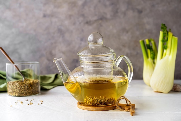 Thé au fenouil dans une théière en verre, bulbe de fenouil, graines sur table en bois blanc et fond gris