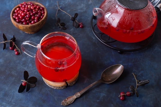 Thé aux canneberges chaud dans une tasse en verre et une théière sur un fond sombre et froid.