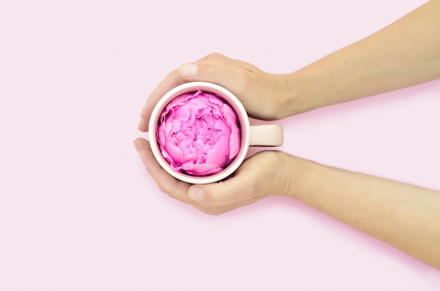 Thé aux fleurs d'été dans les mains des femmes. tasse créative avec pivoine sur fond rose.