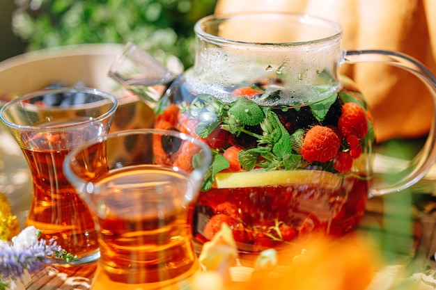 Thé aux fruits aromatiques naturels dans une théière transparente et dans une tasse parmi les baies et les fleurs. thé aromatique réchauffant avec un arôme profond de baies et de fleurs sauvages.